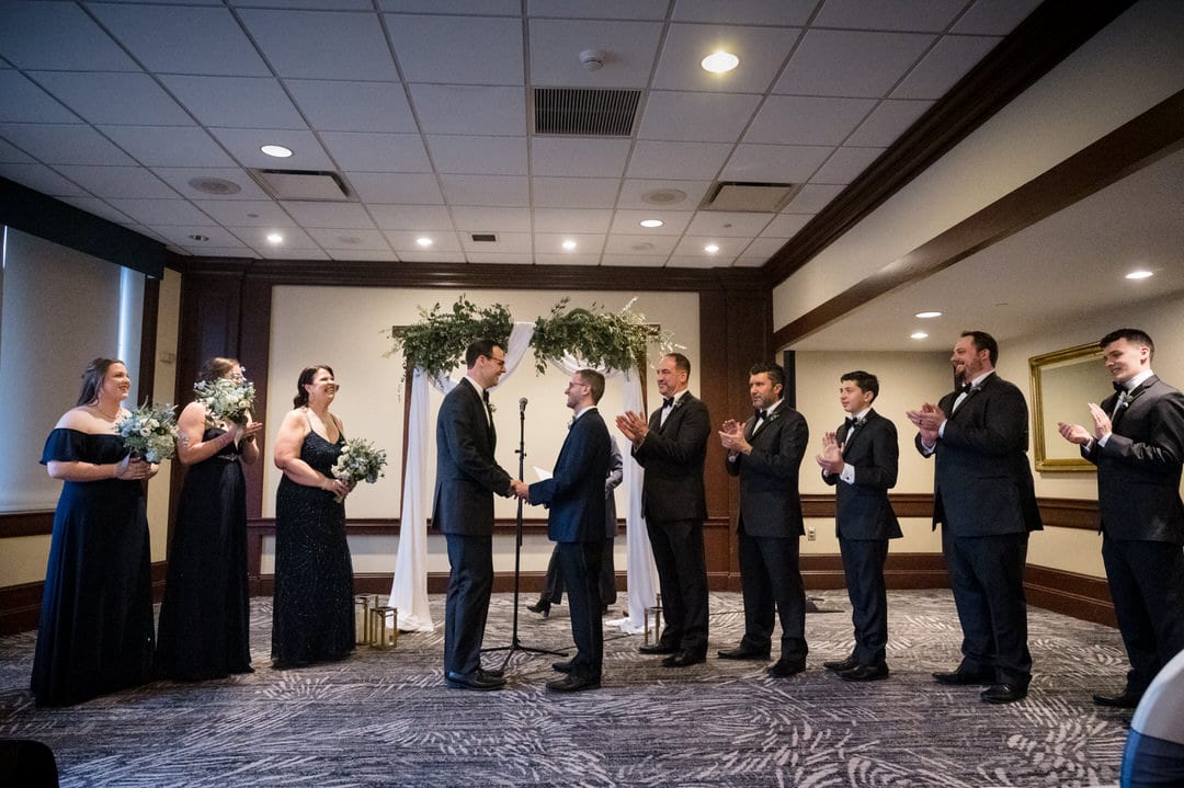 Two grooms holding hands as they exchange rings with their wedding party arrayed on either side of them.