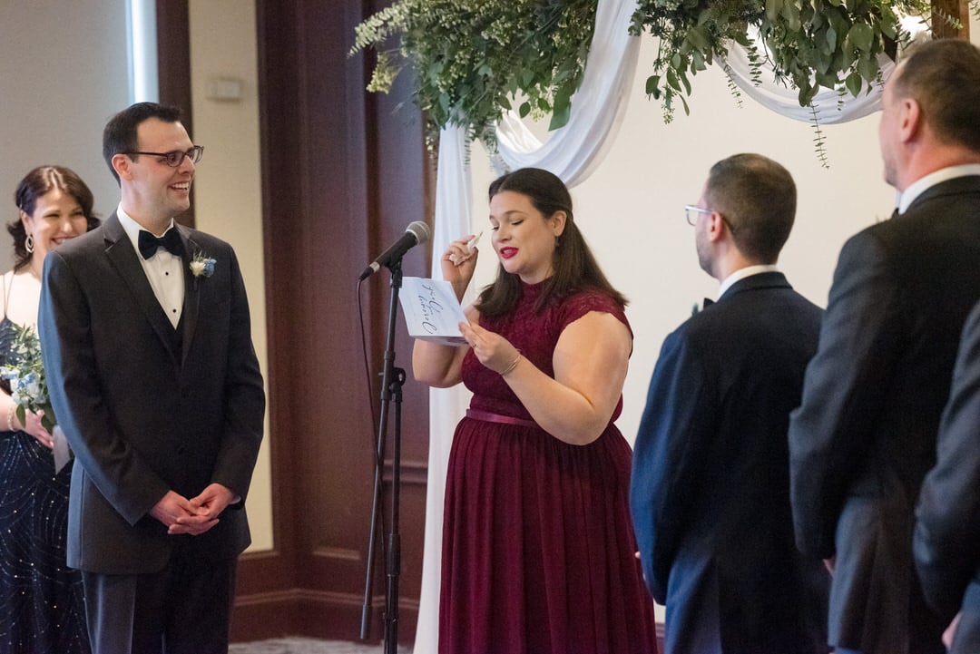 A woman in a dark red dress wipes a tear away as she reads during a wedding ceremony for two grooms.