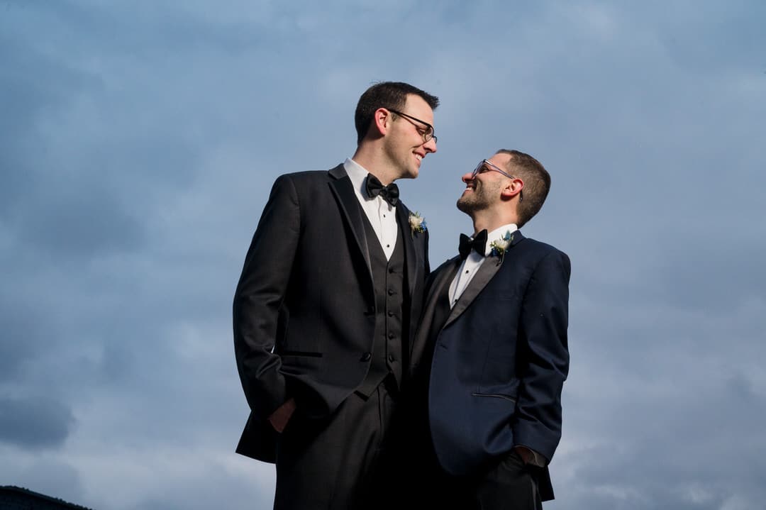 Two grooms look at each other standing in front of a cloudy sky.