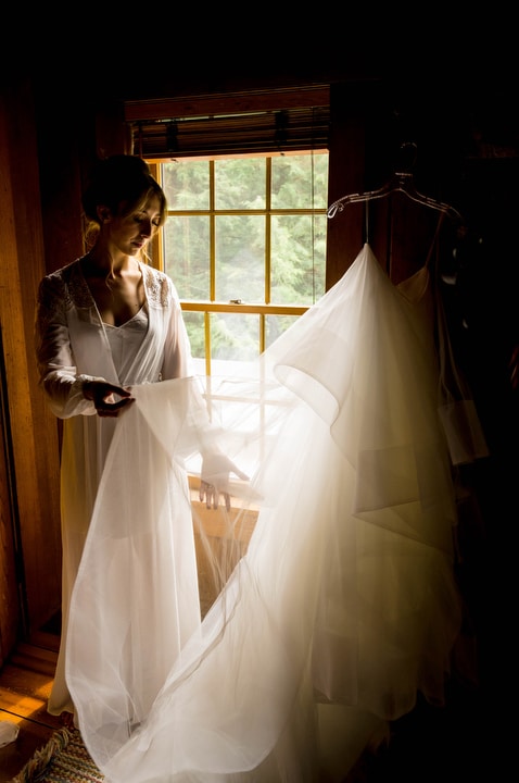 A bride stands in front of a window as she holds a sheer wedding veil.