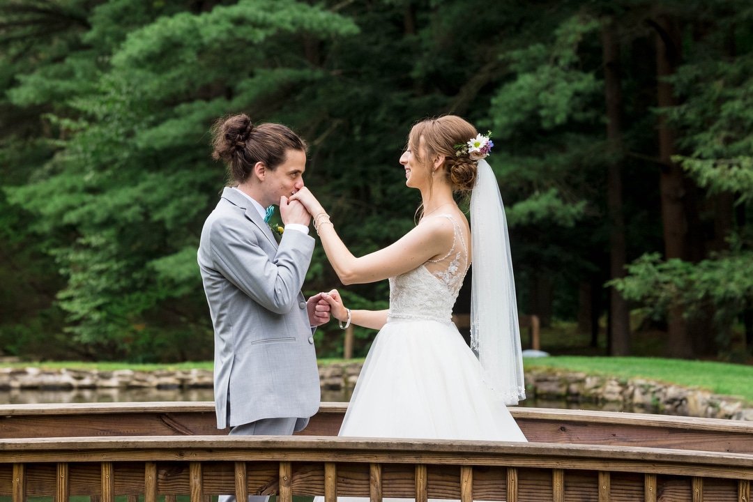 A groom with a ponytail kisses his bride's hand as they stand on a bridge during a summer wedding at seven springs.