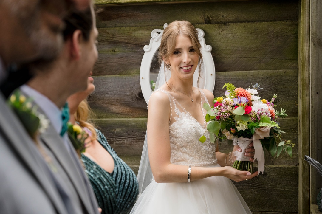 A bride holds her flowers and looks over her right shoulder at her groom as he and his parents prepare to walk down the aisle.