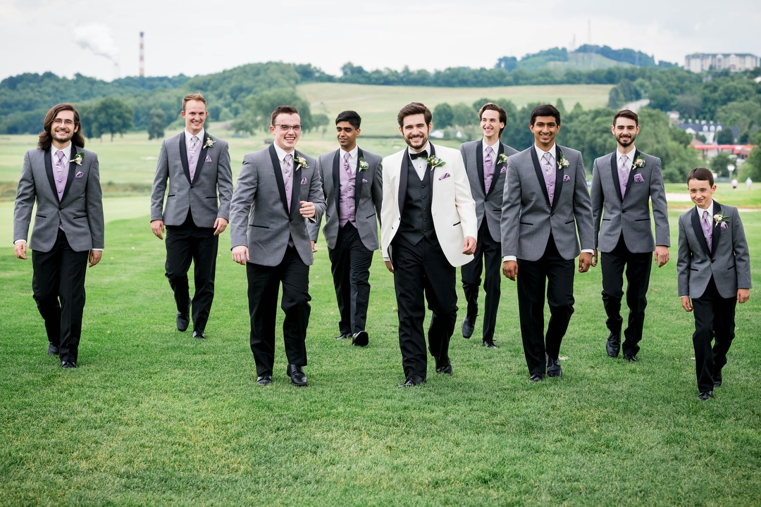 Groomsmen in gray tuxedos accompany a groom wearing a white jacket as they walk near the first tee at Oakmont Country Club before his wedding.