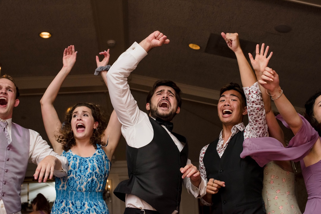 A groom raises his hands in the air while dancing along with wedding guests.