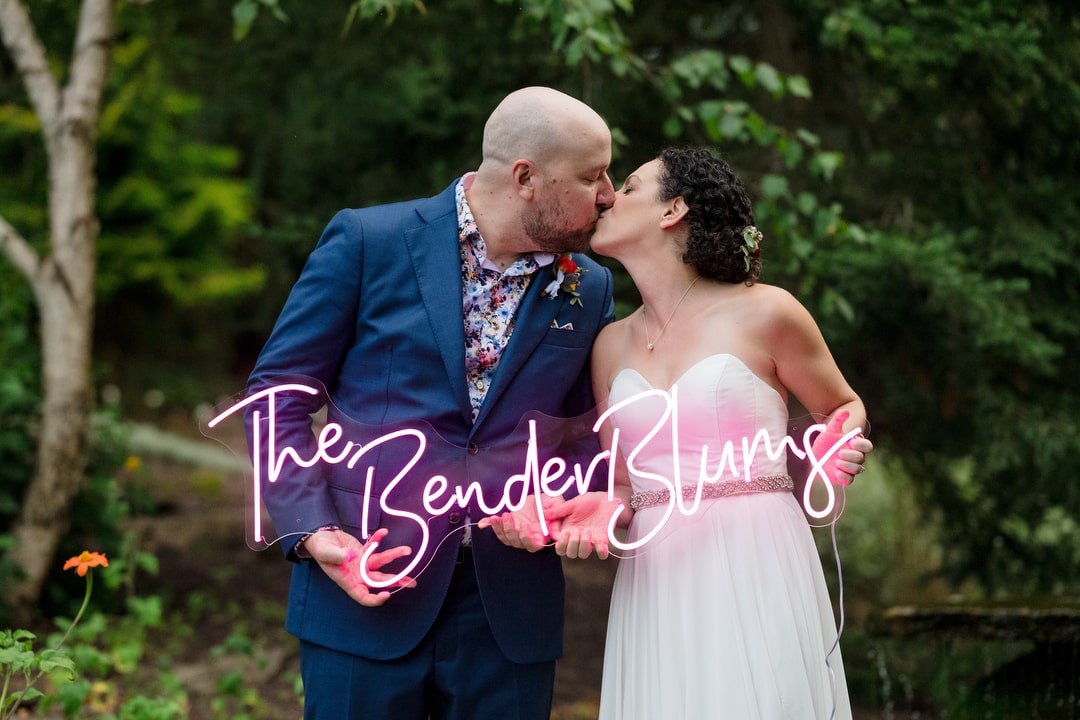 A bride and groom kiss as they hold a pink neon sign during their wedding at the Gardens of Stonebridge