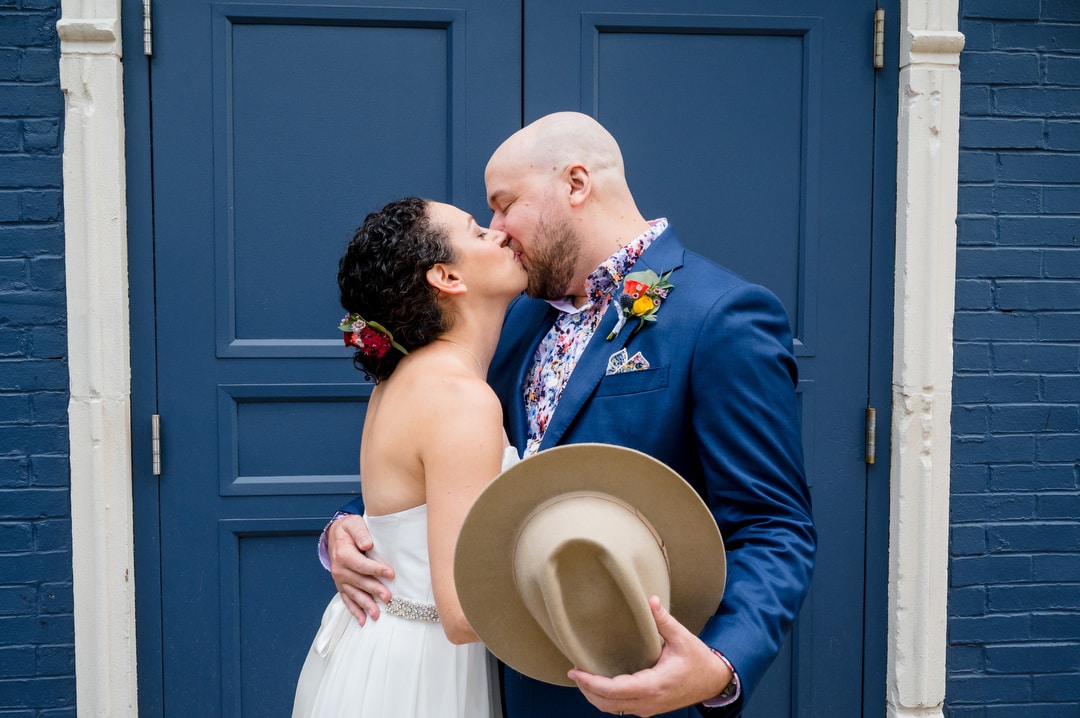 A groom holds his hat as he and his bride kiss in front of a set of blue doors at the Hotel Indigo in Pittsburgh.