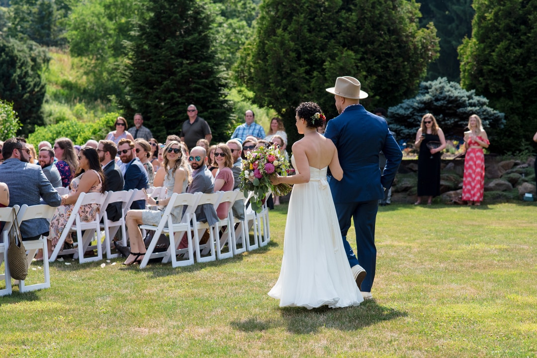 A bride and groom walk down the aisle at their outdoor wedding as their guests look on from their white folding chairs.