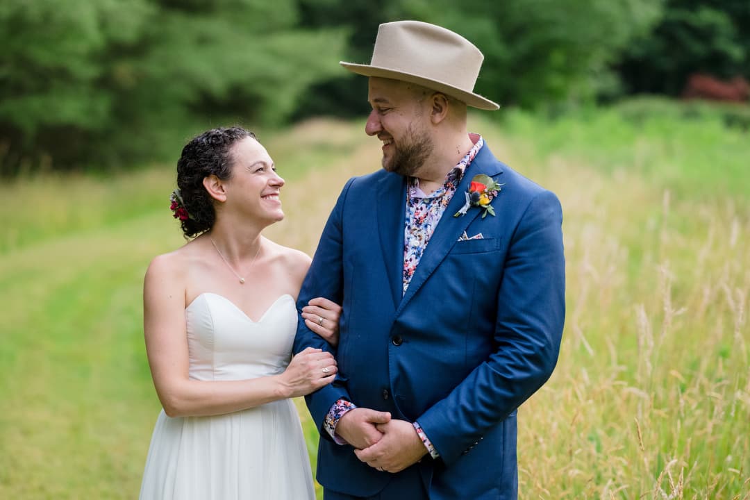 A groom wearing a hat smiles at his bride as they stand in a field at the Gardens of Stonebridge.