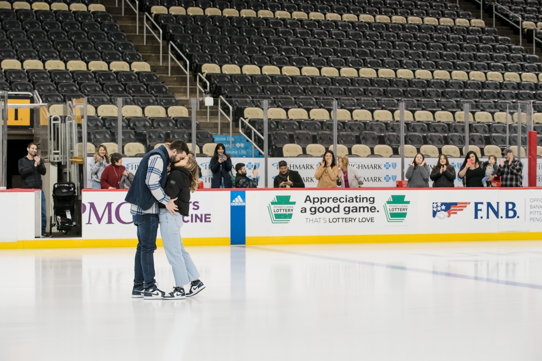 A couple kisses while standing on the ice at PPG Paints Arena in Pittsburgh after a wedding proposal. Their families applaud in the background.