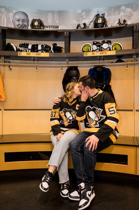 A couple sits and kisses in the Pittsburgh Penguins' locker room at PPG Paints Arena wearing Pens jerseys after their wedding proposal.