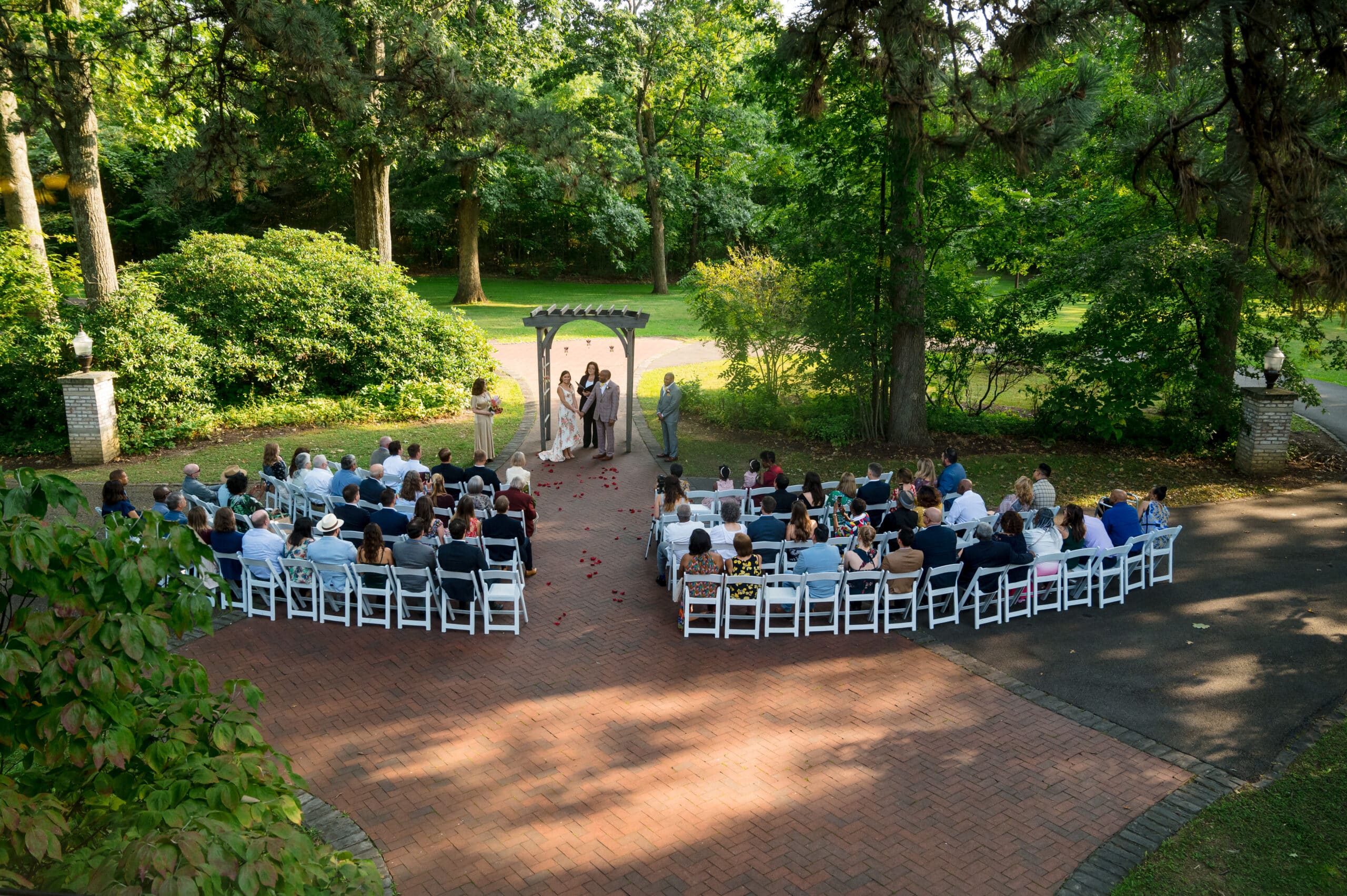 Seen from above, a bride and groom exchange vows under an arch as their guests sit on white folding chairs at Succop Nature Park