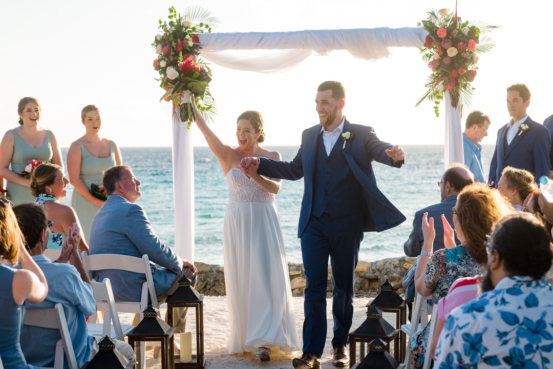A couple raises their hands as they walk down the aisle together after their destination wedding on a beach in Aruba.