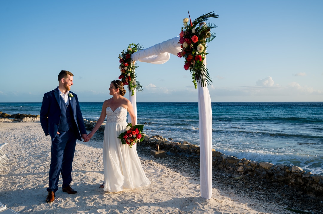 A bride and groom hold hands by an arbor on a beach in Aruba after their destination wedding.
