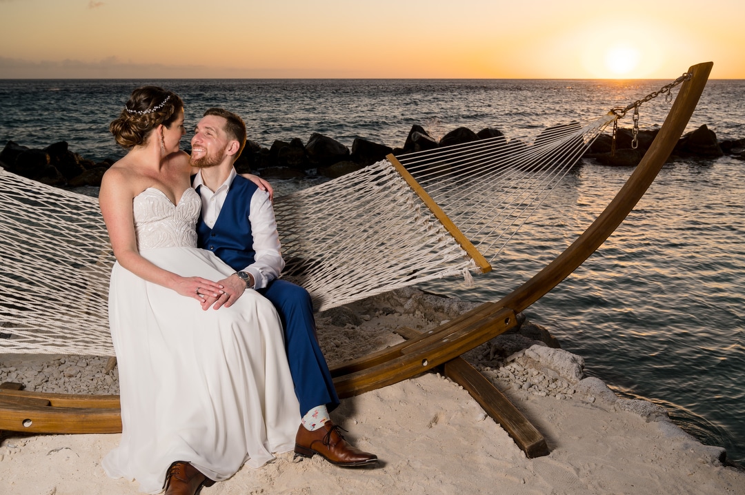 A bride and groom sit together on a hammock at sunset on a beach after their wedding in Aruba.