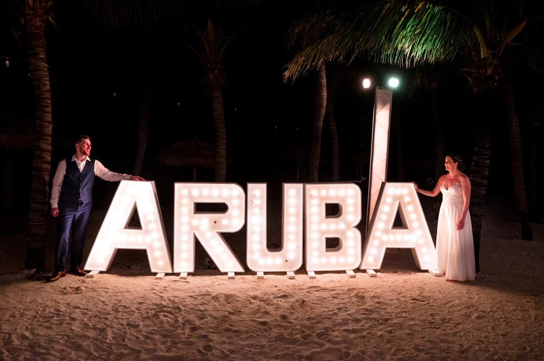A bride and groom stand on opposite ends of a lighted ARUBA sign at their destination wedding.