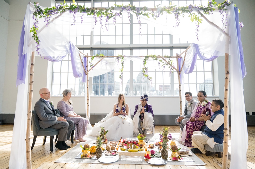 A couple sits under a mandap in front of the large windows at the Energy Innovation Center during their wedding.