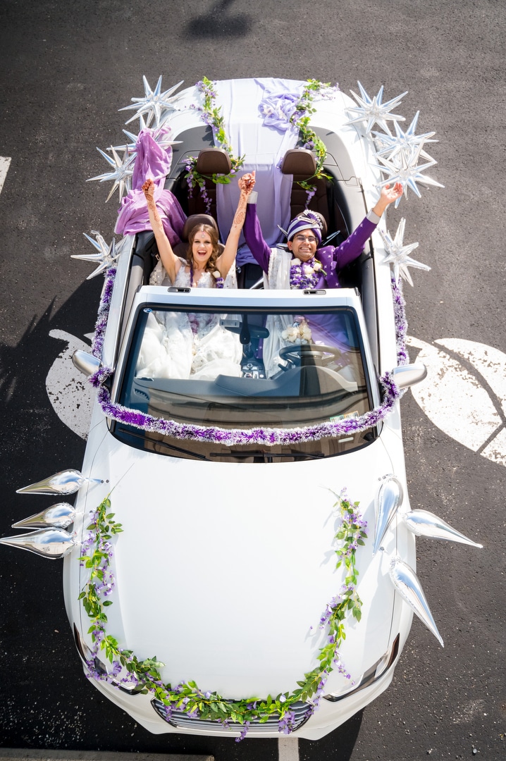 Seen from above a couple seated in an Audi convertible raise their arms in celebration after their Energy Innovation Center wedding.