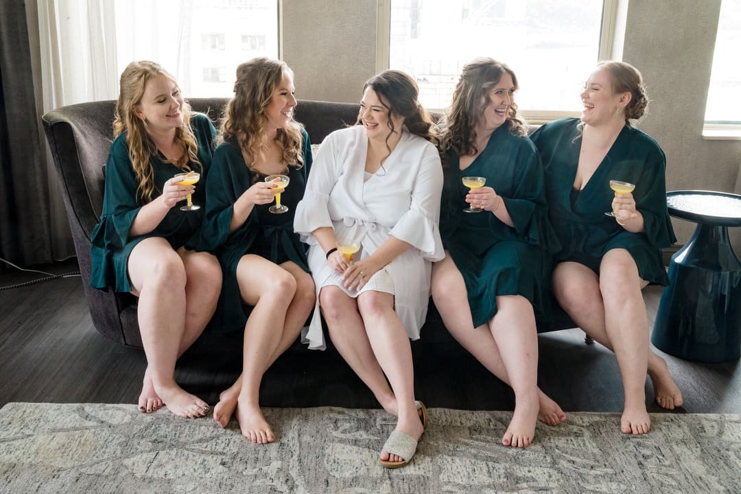 A bride sits on a couch with her bridesmaids as they wear emerald green dressing gowns.