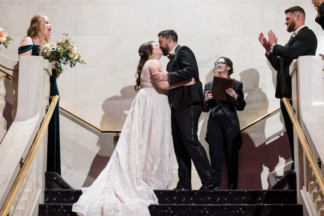 A bride and groom share a kiss on the landing of the grand staircase to the applause of their bridal party.