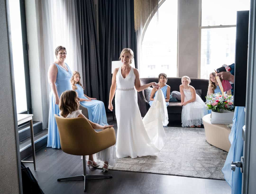 A bride smiles as she stands in her suite at the Pittsburgh Renaissance hotel. Bridesmaids wearing blue dresses surround her.