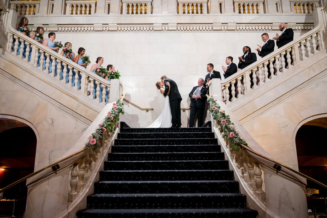 A bride and groom kiss at the end of their wedding ceremony on the stairs of the Pittsburgh Renaissance hotel. Their bridal party applauds.