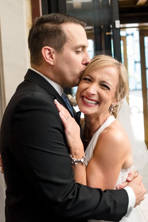 A bride smiles as she embraces her groom. He kisses the side of her head as he holds her in the lobby of the Pittsburgh Renaissance Hotel after their wedding.