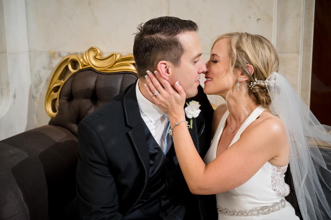 A bride holds her hand on the cheek of her husband as they kiss while seated on a couch in the lobby of the Renaissance Pittsburgh hotel after their wedding.