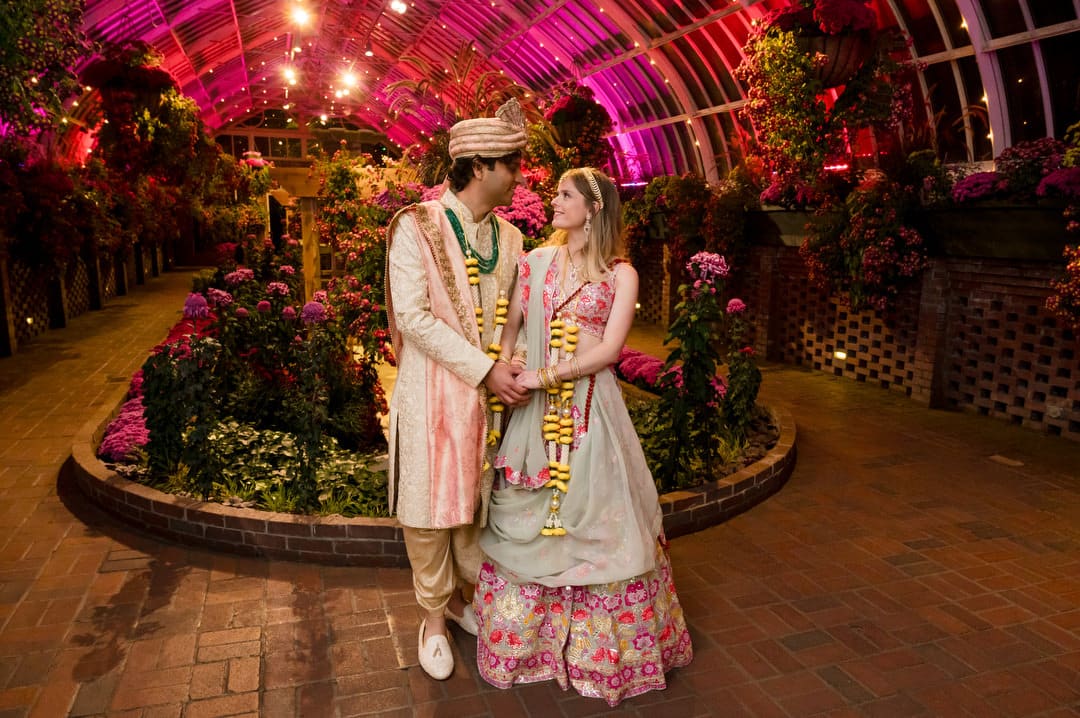 A bride and groom in their south asian wedding garb pose for a photo under the pink lights in the Sunken Garden room at Phipps after their intimate wedding.