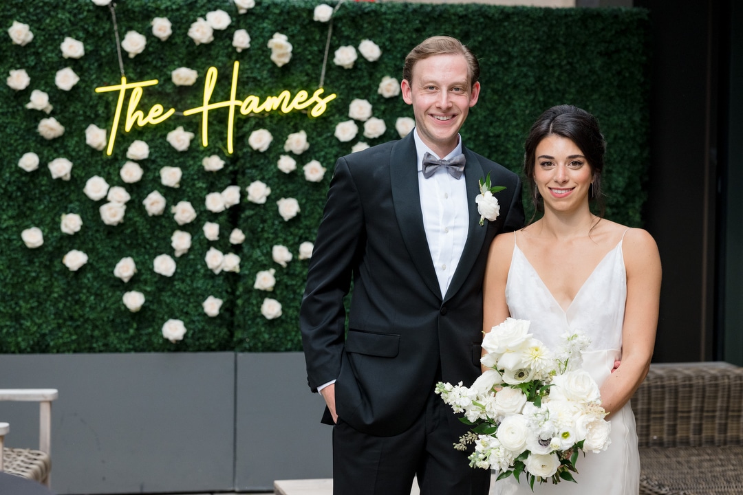 A bride and groom stand together in front of a boxwood wall with a neon sign with their last name on it.
