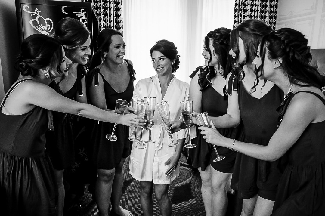 A bride and bridesmaids toast with champagne while wearing dressing gowns in a hotel room.
