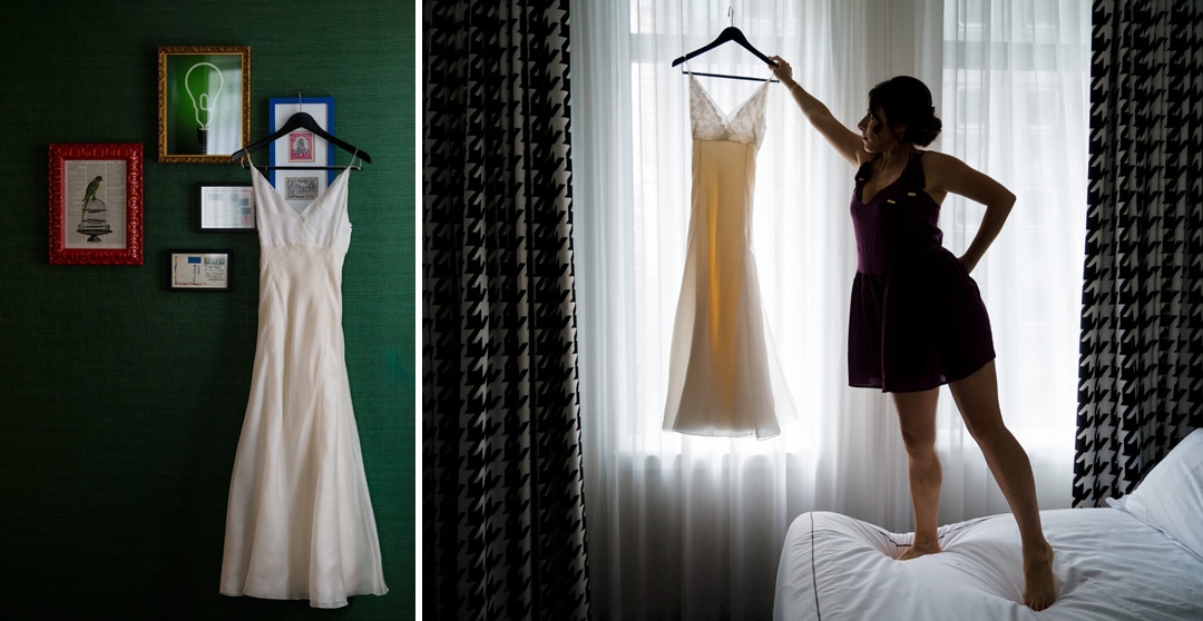 A woman stands on a bed while she holds a dress on a hanger up to a window in a hotel room.