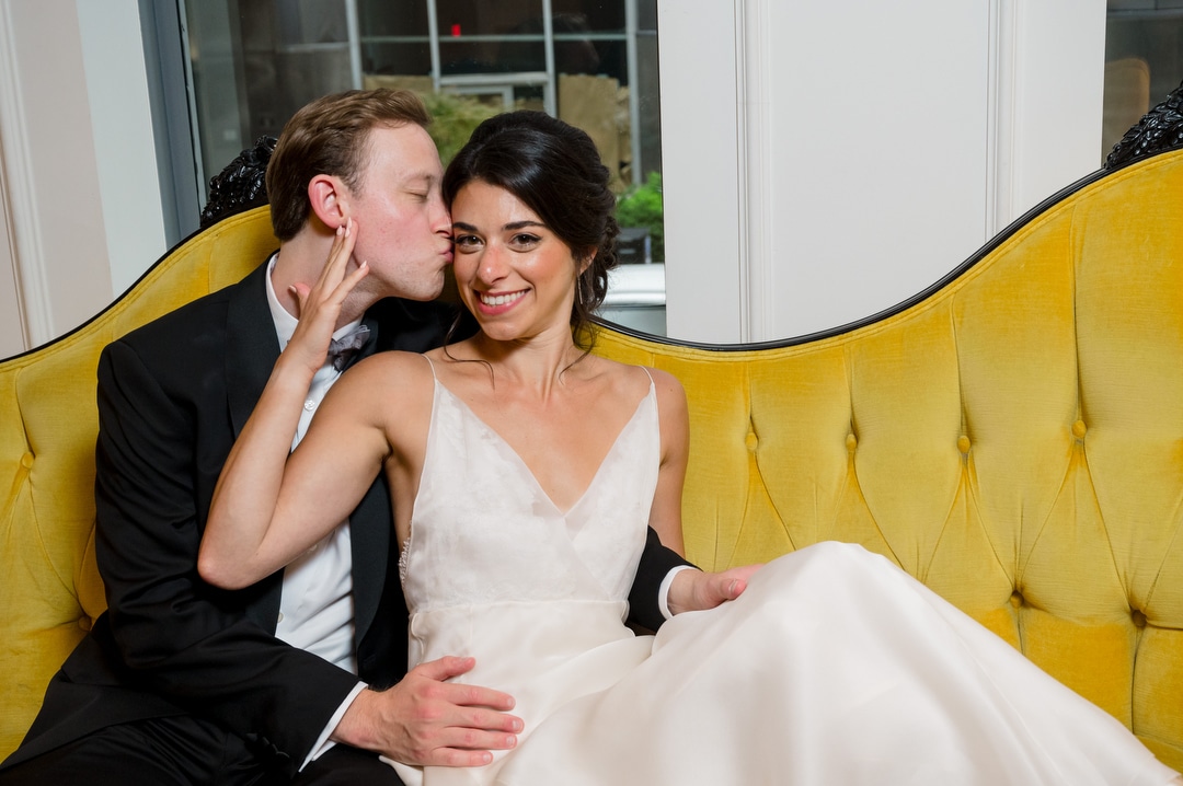 A groom kisses his bride on the cheek as she smiles while they're seated on a yellow couch at the Hotel Monaco in Pittsburgh.