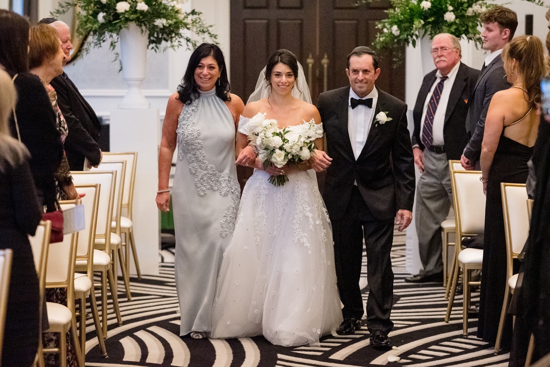 A bride is walked down the aisle by her parents in elegant attire during her wedding at the Hotel Monaco in Pittsburgh.