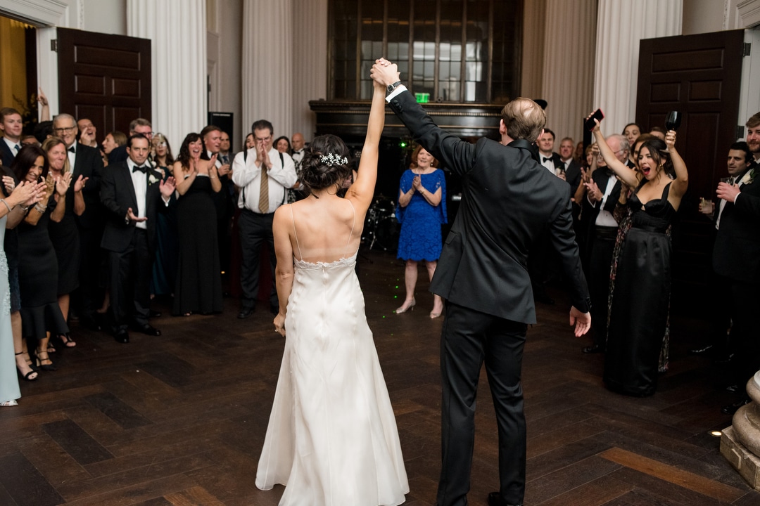 A bride and groom hold their clasped hands into the air as their guests applaud after their first dance.