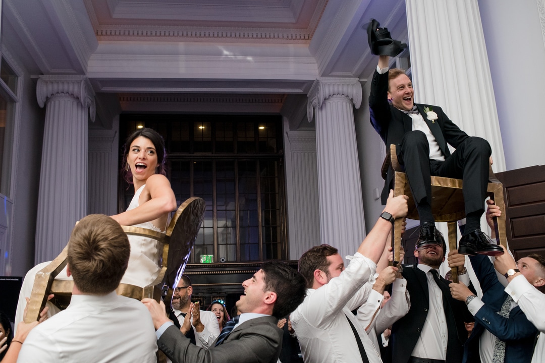 A bride and groom laugh as they are lifted up on chairs in a room with large white columns during their elegant wedding at the Hotel Monaco in Pittsburgh.