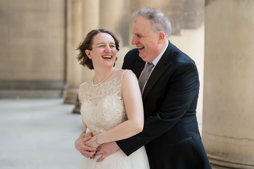 A bride and groom smile and embrace at the columns of the Mellon Institute in Pittsburgh after their wedding.