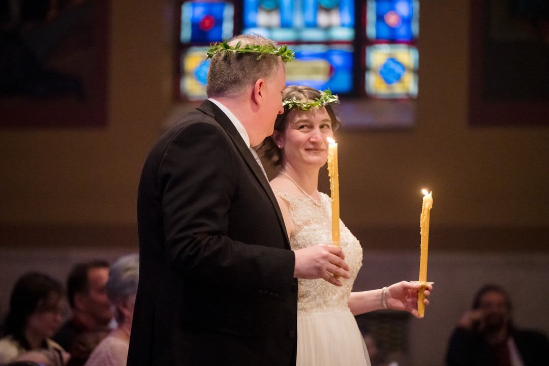 A bride and groom wearing laurel crowns and holding long taper candles look at each other and smile during their wedding ceremony at Saint John Chrysostom church in Pittsburgh.