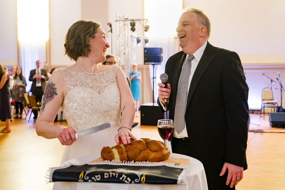 A bride and groom look at each other and laugh as they cut a loaf of challah bread during their Saint Nicholas Cathedral Room wedding.