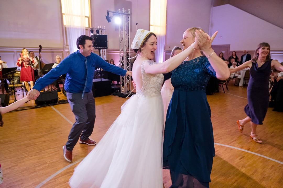 A bride wearing a bonnet dances with a guest as other guests dance in a circle around them at a Saint Nicholas Cathedral Room wedding in Pittsburgh.