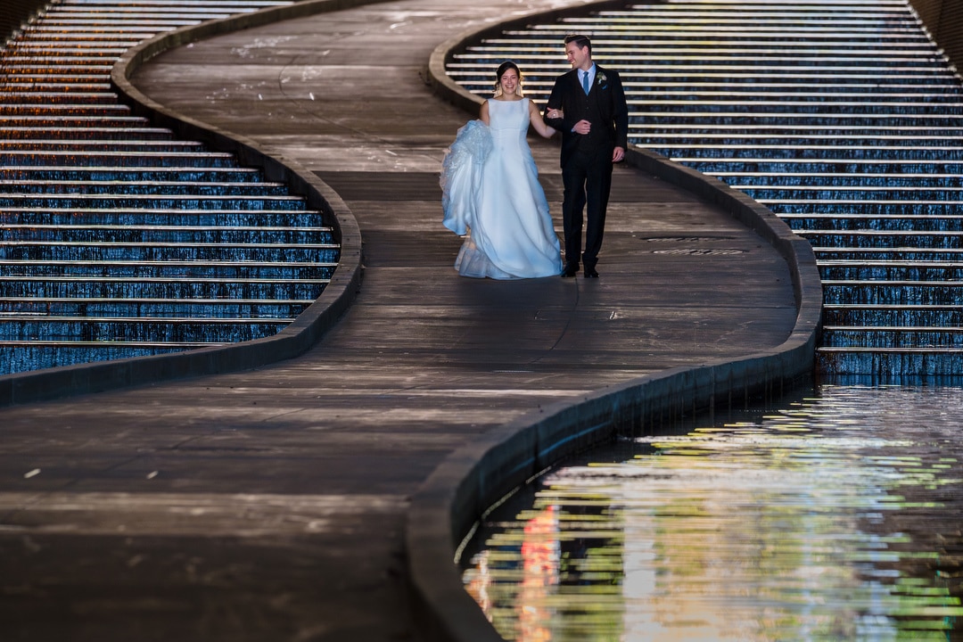 A bride and groom experience The Art Room Wedding as they walk along a curving pathway beside cascading water steps. The bride, in a white gown, and the groom, in a black suit, revel in serenity. Reflections in the water enhance the romantic atmosphere of their special day.