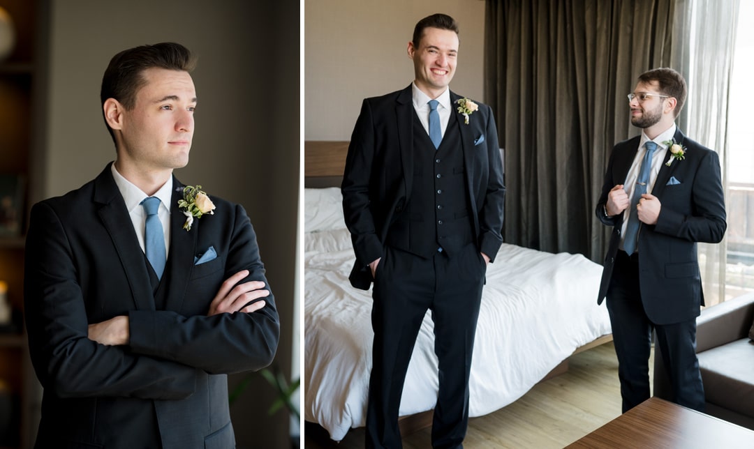 The groom, clad in a black suit with a floral boutonnière, stands with arms crossed, gazing thoughtfully out the window. In another scene from The Art Room Wedding, he chats and smiles with a groomsman in matching attire in a well-lit room near a bed.