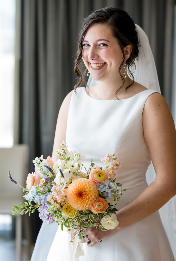 A smiling bride in a white dress, from The Art Room Wedding collection, holds a colorful bouquet with orange, pink, and purple flowers. She stands indoors against a curtain backdrop, her hair styled in loose curls beneath her veil.