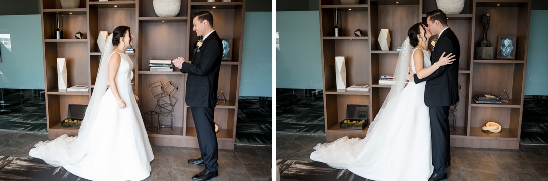 In The Art Room Wedding, a bride and groom stand facing each other in a modern space, their joy evident. The bride in her white gown and the groom in a dark suit hold hands, embracing warmly before a wooden shelf adorned with books and decor.