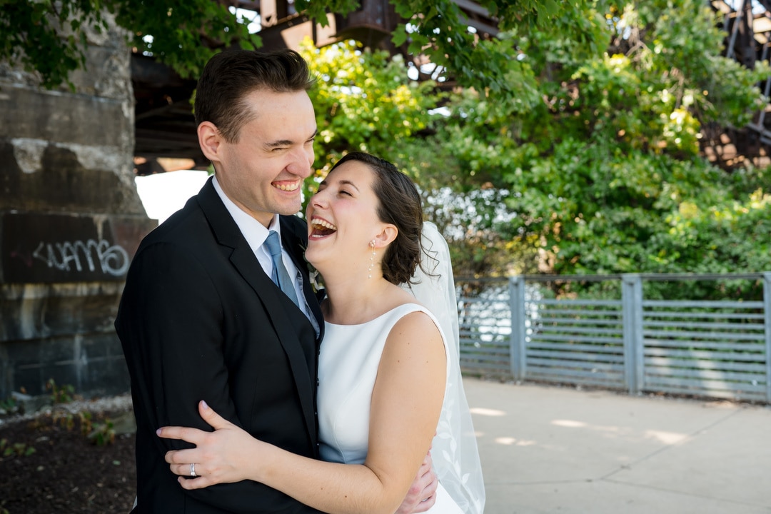 A joyful couple in wedding attire from The Art Room Wedding embraces and laughs under a leafy tree beside a metal bridge. In the background, graffiti adorns a concrete wall and metal railing, while sunlight filters through the leaves.