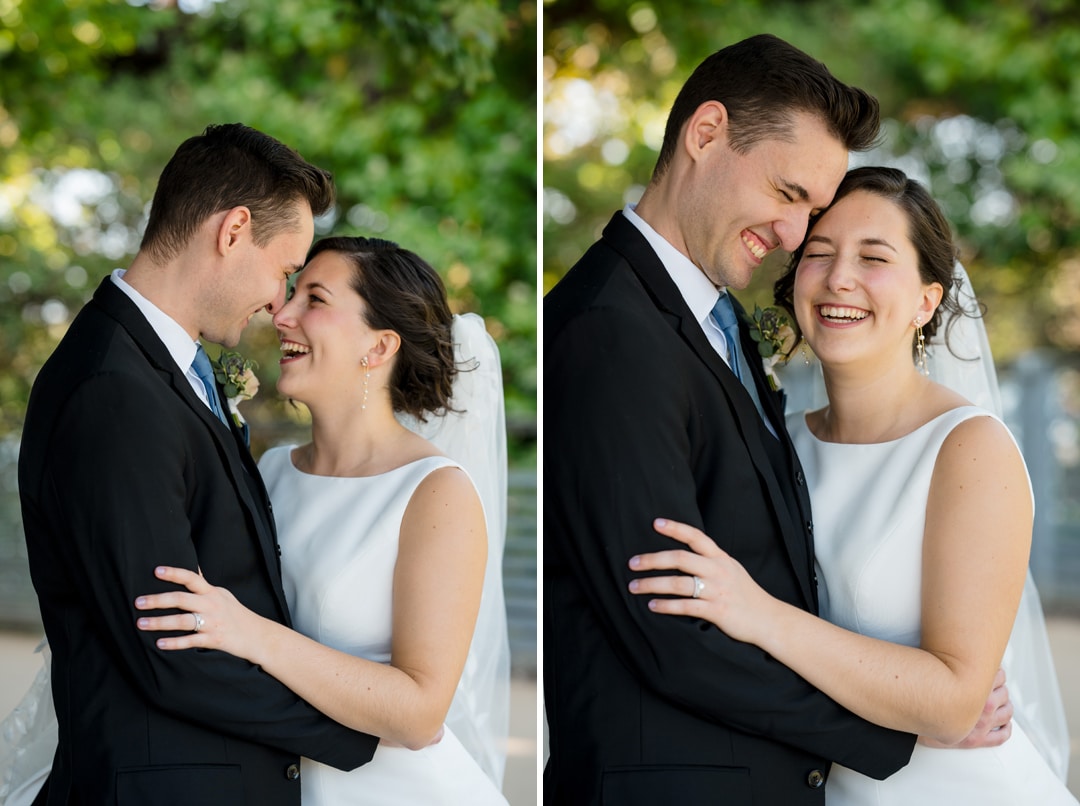 A bride and groom stand closely, smiling and embracing on their special day at The Art Room Wedding. The bride glows in a white dress and veil, while the groom looks dapper in a black suit and tie. They are outdoors with green foliage as their backdrop, radiating joy and happiness.