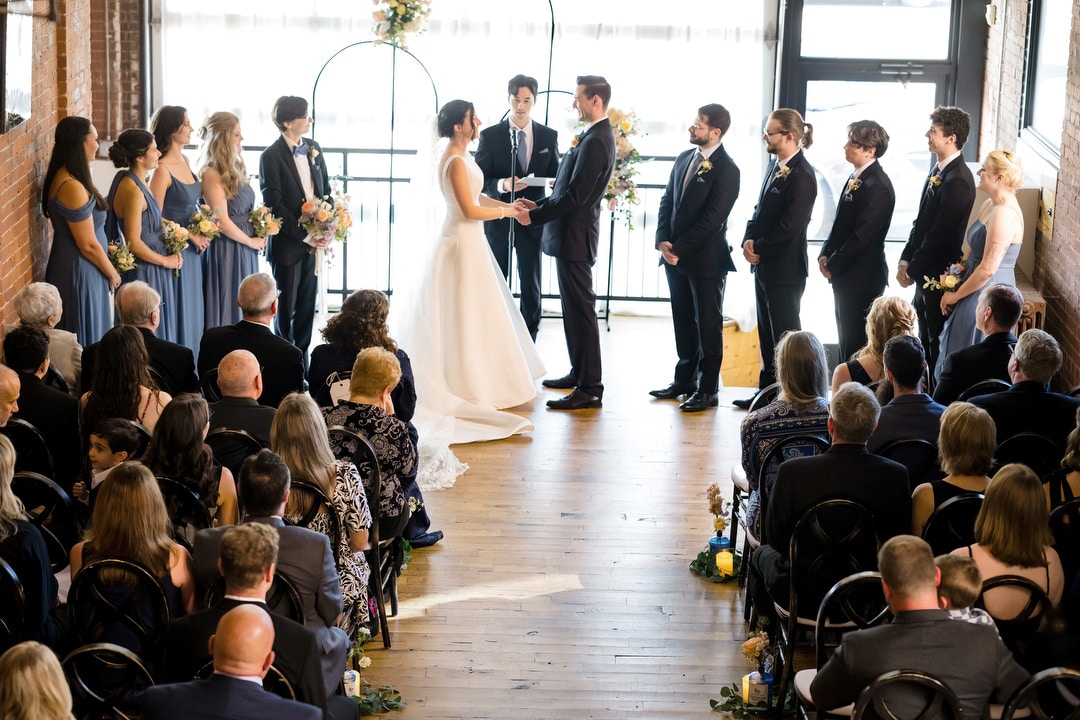 The Art Room Wedding features a bride and groom holding hands at the altar, surrounded by bridesmaids in blue dresses and groomsmen in black suits. Guests are seated on both sides in this bright, decorated venue with large windows, creating a picture-perfect ceremony.