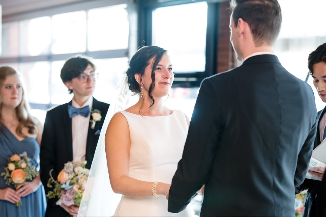 The Art Room Wedding is in full swing as the bride and groom hold hands, their smiles reflecting the joy of the moment. Surrounded by bridesmaids and groomsmen with bouquets, they stand bathed in natural light streaming through large windows, capturing this beautiful indoor ceremony.