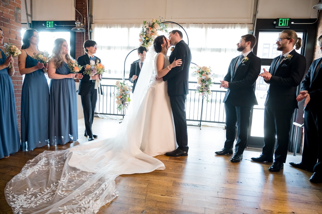 At The Art Room Wedding, a bride and groom share a kiss amidst bridesmaids in blue dresses and groomsmen in dark suits. The brides long white veil elegantly flows on the wooden floor, while exquisite floral arrangements adorn the setting.