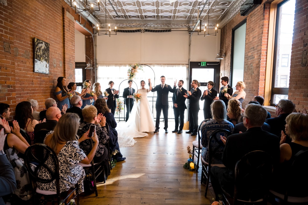 A newlywed couple stands at the altar in The Art Room Wedding, a rustic brick-walled venue, surrounded by their wedding party. Guests are seated on either side, capturing the moment with applause as soft natural light filters through large windows.