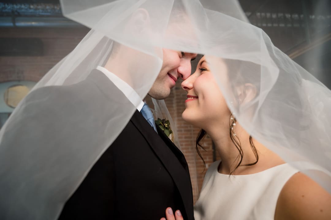 A bride and groom share a joyful moment under a veil at The Art Room Wedding. The bride, in a white dress, and the groom, dressed in a dark suit with a boutonniere, smile at each other while standing in front of a rustic brick wall.